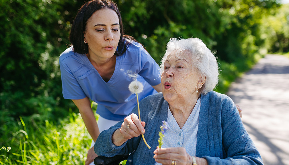 Photo of Caregiver Blowing Dandelion With Elderly Woman